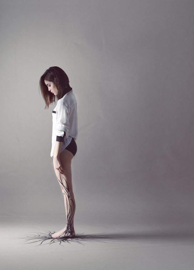 Mariana Esquivel's artistic portrayal of a female figure with roots emerging from her legs set against a minimalist gray backdrop.